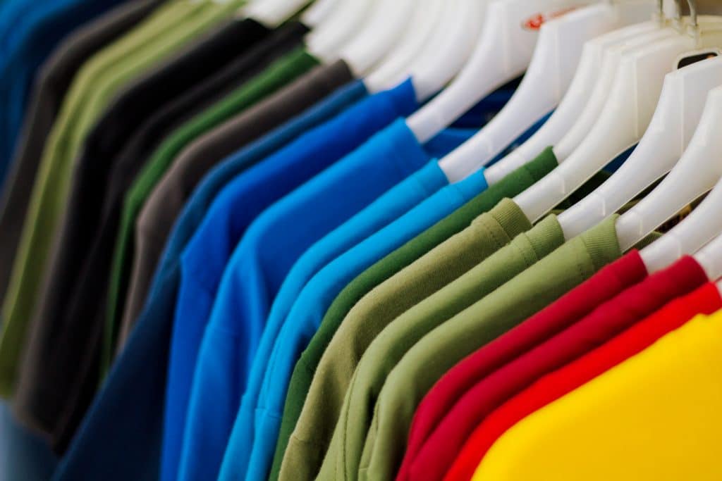 A row of colorful t-shirts, including blue, green, red, yellow, and gray, hangs neatly on white plastic hangers on a clothing rack—kept spotless with commercial lint rollers.