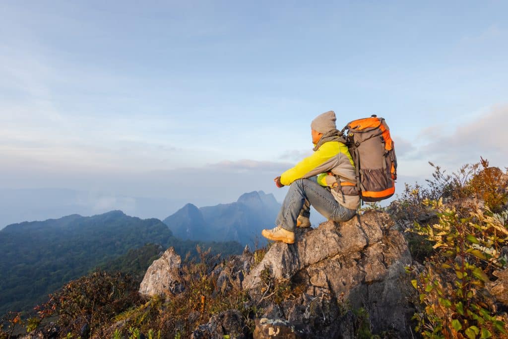 A hiker wearing a yellow jacket, gray beanie, and backpack sits on a rocky ledge overlooking a scenic mountain range under a clear sky, bulk lint rollers packed for easy clean-up among the autumn foliage.