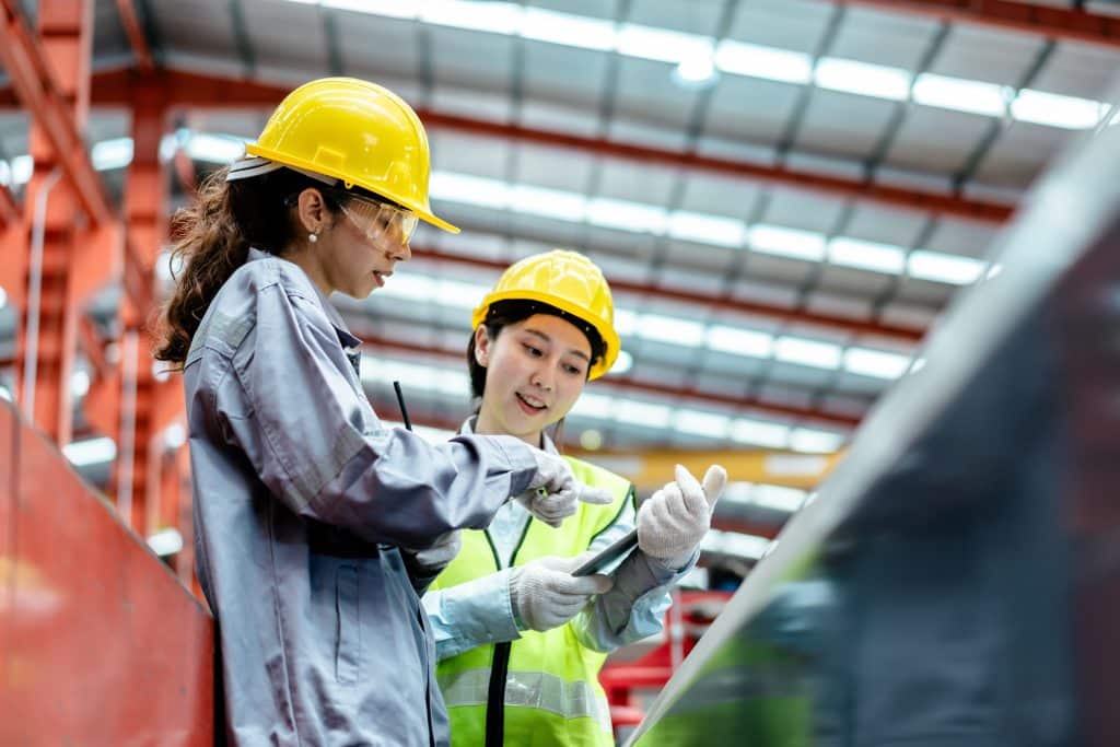 Two women wearing hard hats, safety glasses, and reflective vests are discussing something on a tablet inside a large manufacturing facility, possibly reviewing the use of industrial lint rollers for workplace cleanliness.