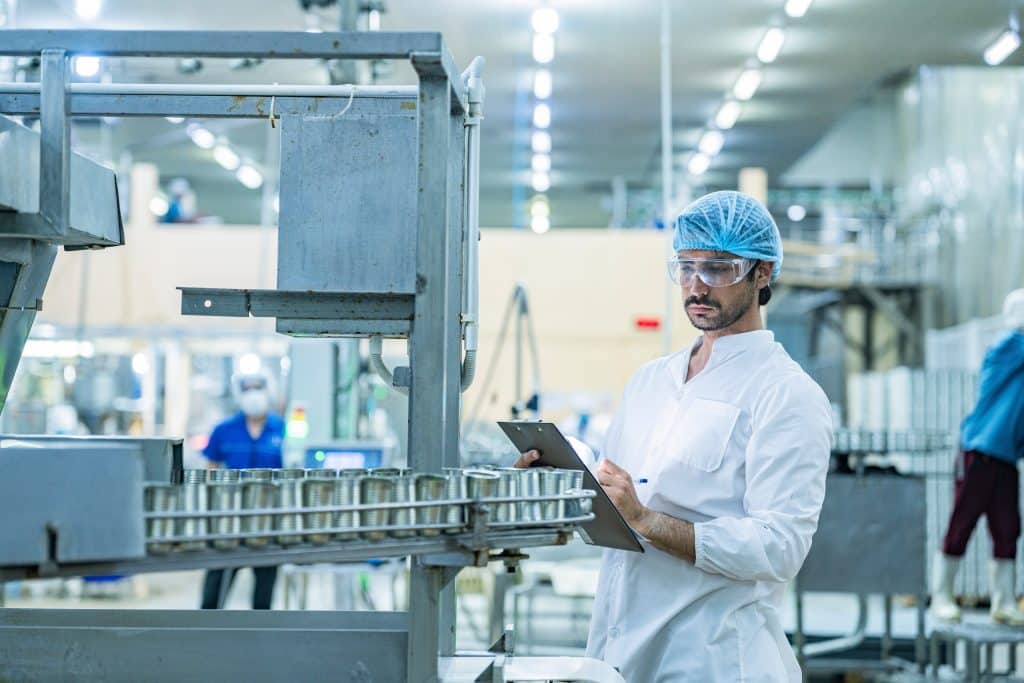 A factory worker wearing safety glasses, a hairnet, and a white lab coat inspects machinery and takes notes on a clipboard, ensuring cleanliness with industrial lint rollers in a brightly lit industrial setting.