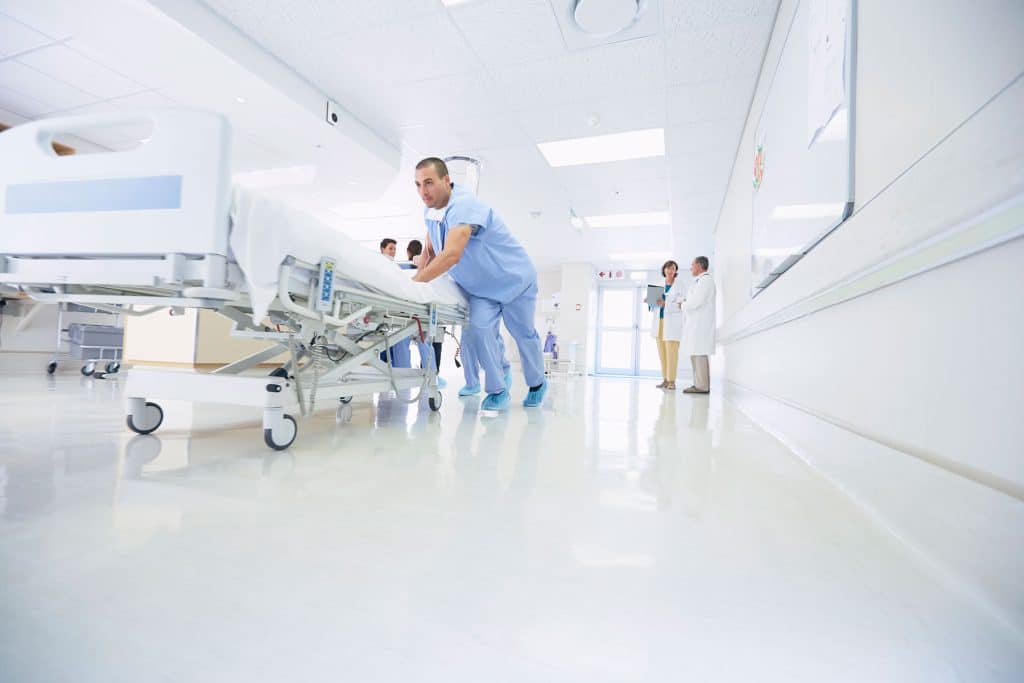 A healthcare worker in scrubs, equipped with bulk lint rollers, pushes an empty hospital bed down a bright, clean hallway, while three medical professionals stand together in the background.