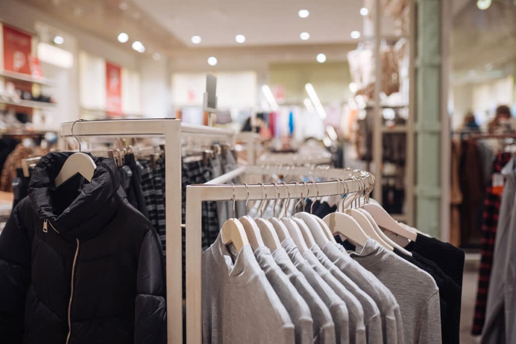 Clothing store interior with racks of neatly hung gray sweaters and a black puffer jacket in the foreground, and various other clothes displayed throughout the bright, modern store.
