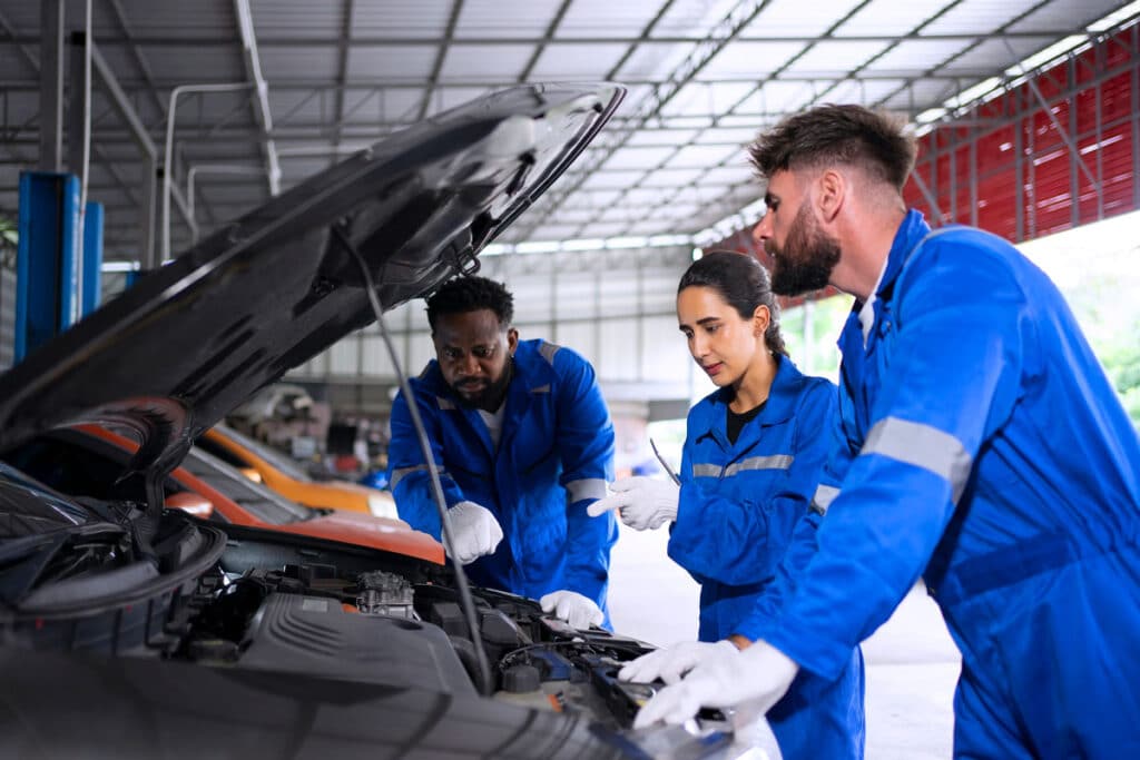 Three auto mechanics in blue uniforms examine and discuss a car engine under the hood in a well-lit garage. Two men and one woman focus intently on diagnosing the vehicle issue.
