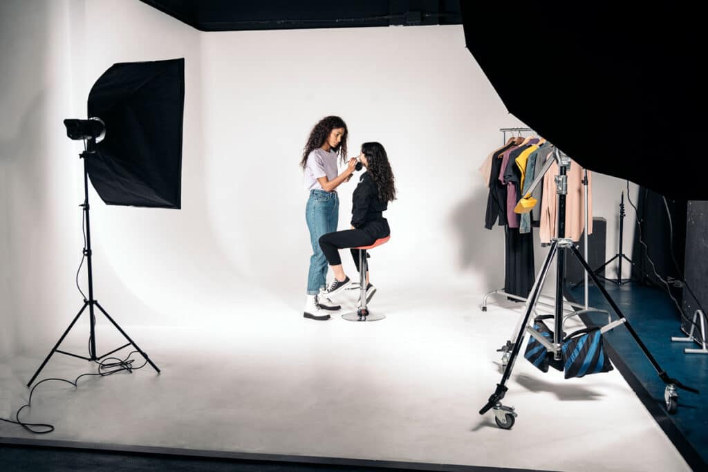 A makeup artist applies makeup to a seated woman in a photography studio with professional lighting, backdrop, and a clothing rack with various outfits in the background.