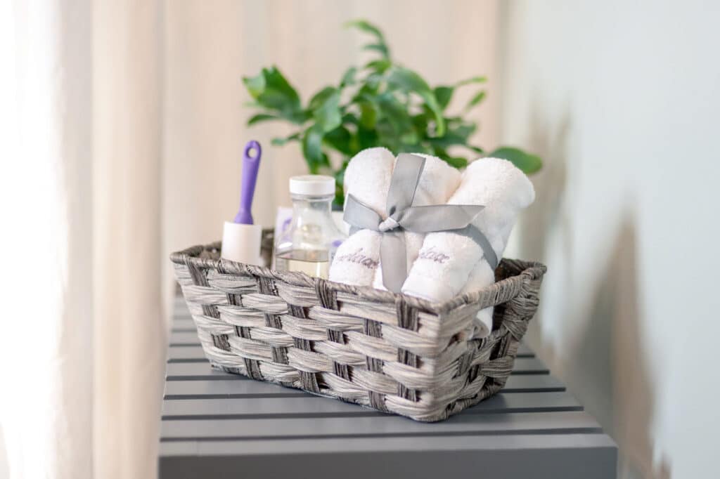 A woven basket on a gray striped surface holds rolled white towels with a gray ribbon, a small bottle, a lint roller with a purple handle, and a green plant in the background.