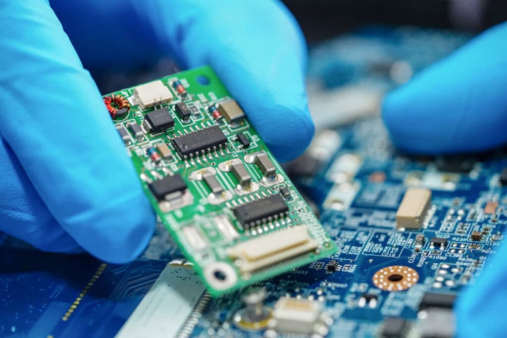 A person wearing blue gloves holds a green printed circuit board above another electronic board, highlighting microchips and components in a close-up, technical setting.