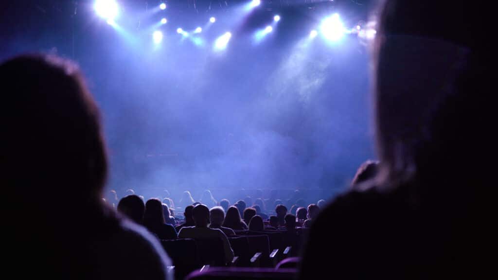Audience members in dark silhouettes sit in rows facing a brightly lit stage, with blue and purple lights and haze creating a dramatic, atmospheric effect.