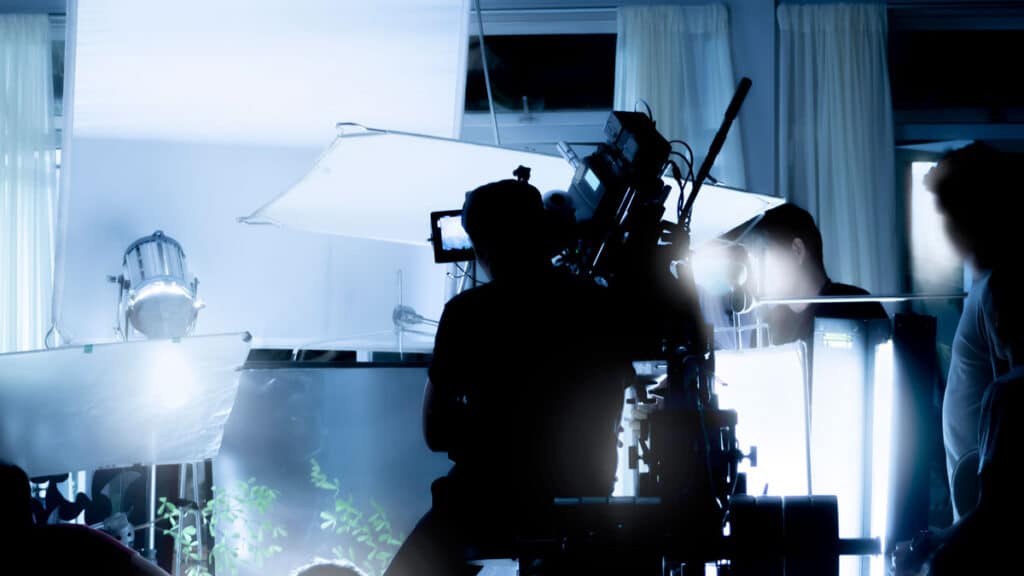 A person operates a large film camera on a movie set, surrounded by bright lights, equipment, and partially visible crew members in a room with white curtains.