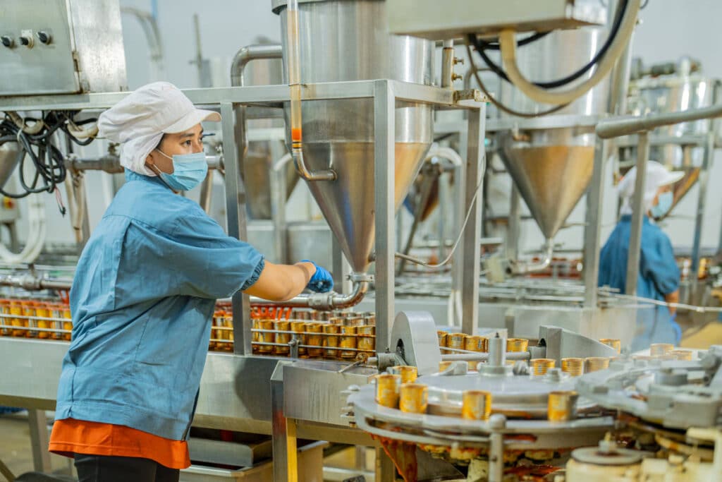 A worker wearing a hairnet, face mask, and gloves operates machinery in a food processing factory, filling containers on a conveyor belt with a liquid or semi-liquid product.