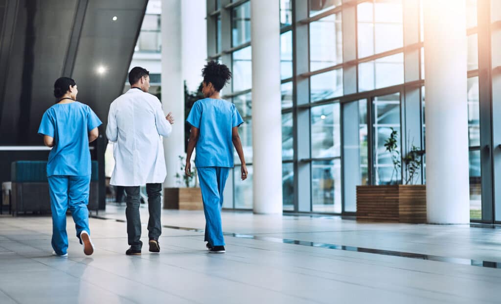 Three medical professionals, two in blue scrubs and one in a white lab coat, walk down a bright hospital hallway with large windows and white columns, viewed from behind.