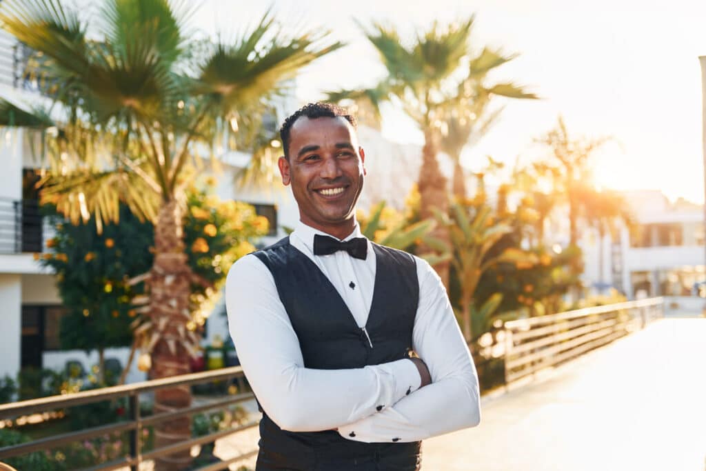 A man wearing a white shirt, black vest, and bow tie stands outdoors with arms crossed, smiling. Palm trees and a sunlit pathway are visible in the background.