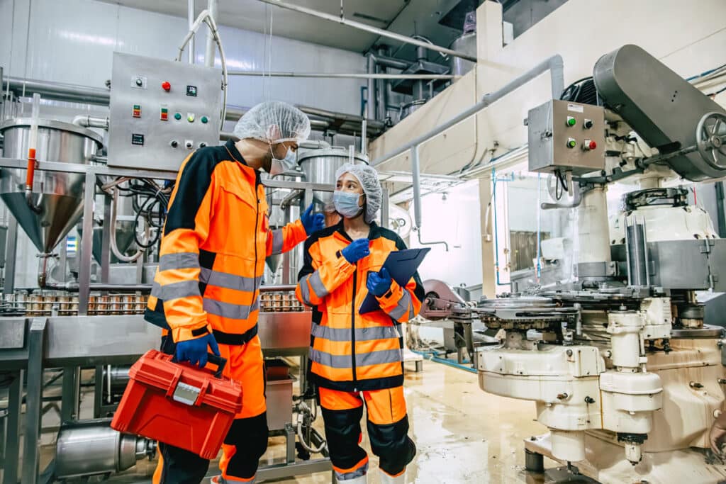 Two workers in orange safety gear, hairnets, masks, and gloves stand in a food processing factory. One holds a red toolbox, the other a clipboard. Industrial machinery surrounds them as they discuss their work.