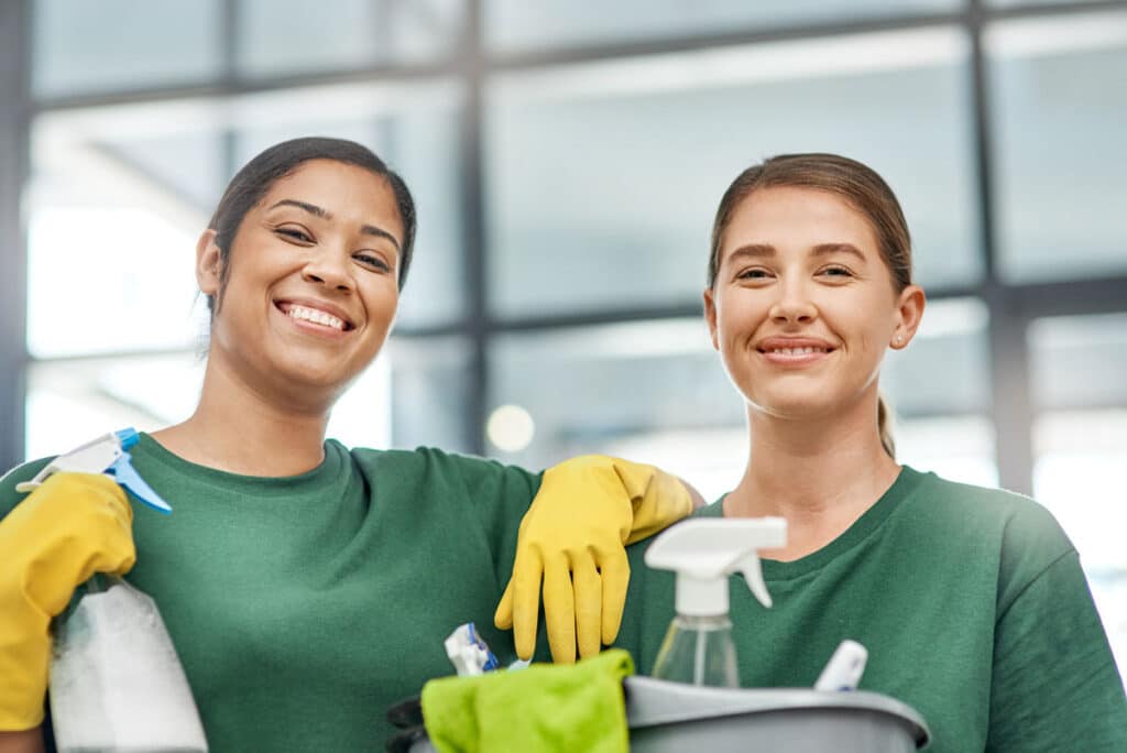 Two women in green shirts and yellow gloves smile while holding cleaning supplies, including spray bottles and a bucket, standing in a bright, modern indoor setting.