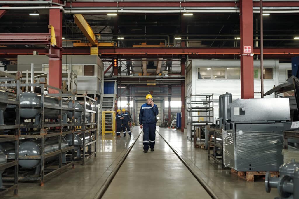 A worker in a yellow hard hat and blue uniform walks down the center aisle of a spacious industrial factory, surrounded by metal equipment, shelves, and other workers in the background.
