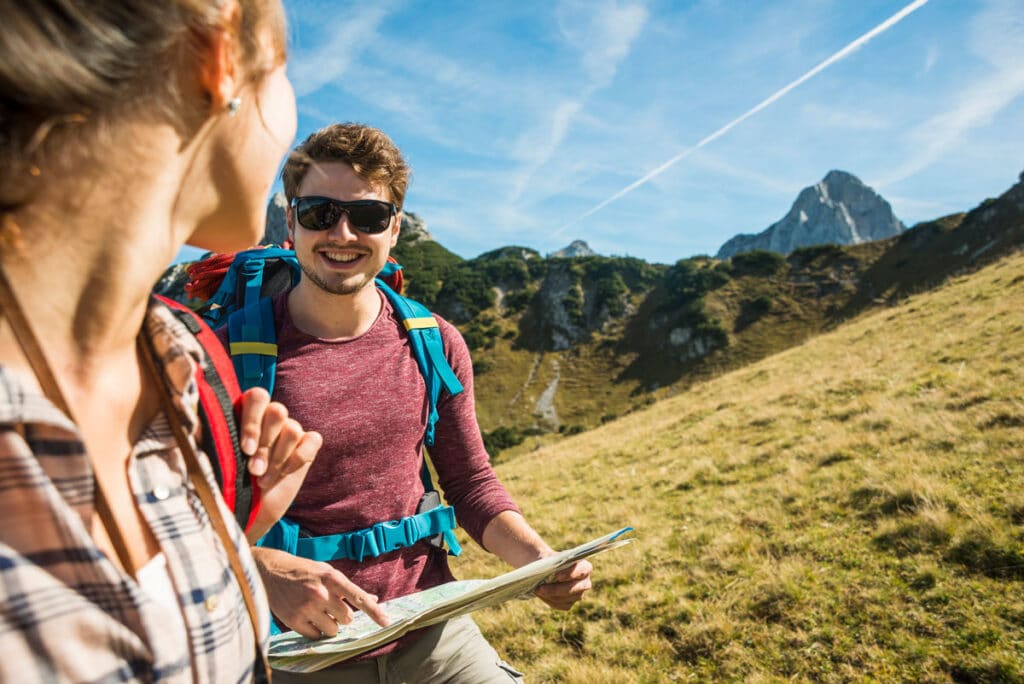 Two young hikers with backpacks smile and look at each other while navigating with a map on a grassy mountain trail under a clear blue sky with distant rocky peaks.