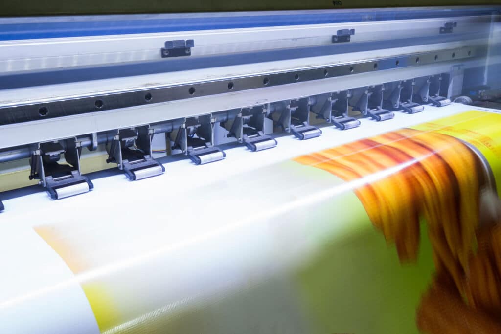 A large-format printer in action, producing a colorful printed banner. The image shows the printer head moving across the material as vivid colors and graphics are being printed.