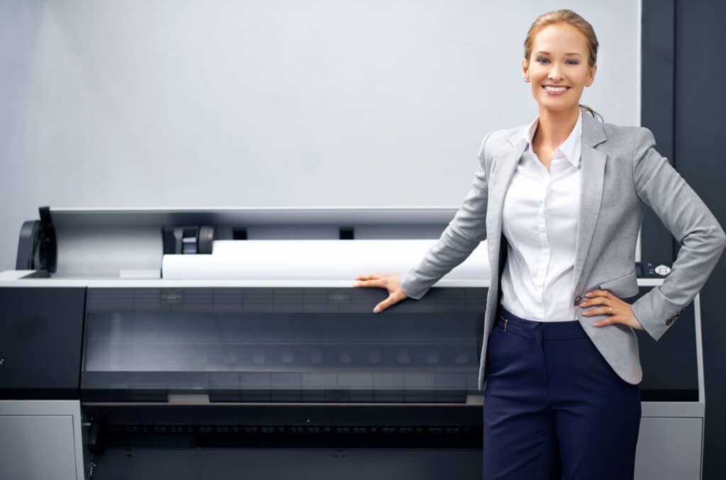 A woman in a gray blazer and white shirt stands smiling with one hand on a large industrial printer in a modern office setting.