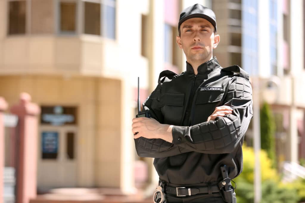 A security guard in a black uniform and cap stands outside a modern building, looking serious with arms crossed and holding a walkie-talkie.