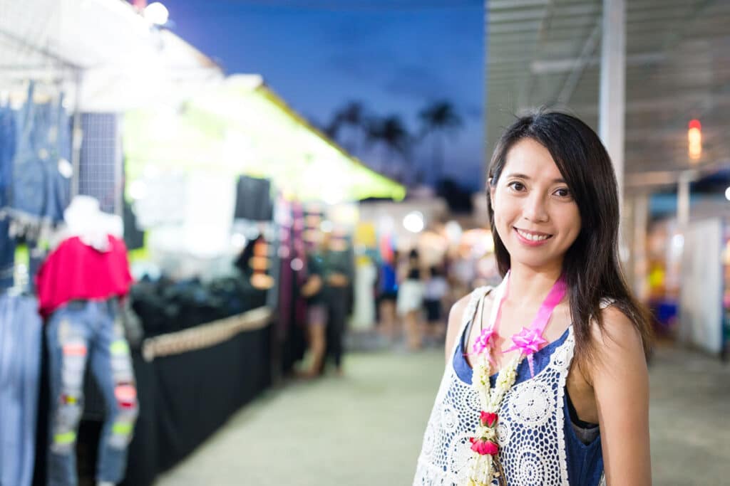 A woman with long dark hair smiles at the camera, wearing a floral lei, at an trade show with bright stalls and mannequins in the background. It is evening, and the event is busy and well-lit.